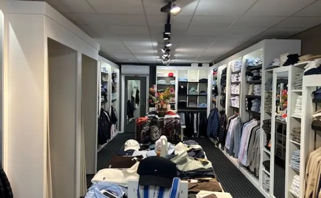 Blik door de winkel met paskamers, kledingrekken en een tafel met petten en truien